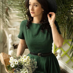 May include: A woman wearing a green dress with a black belt and a black wide-brimmed hat. She is holding a bouquet of white flowers.