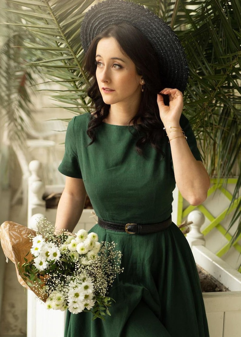 May include: A woman wearing a green dress with a black belt and a black wide-brimmed hat. She is holding a bouquet of white flowers.