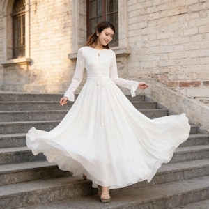 Puede incluir: Un vestido blanco largo con mangas largas y escote de ojo de cerradura. El vestido tiene una cintura entallada y una falda larga que ondea. Las mangas tienen forma de campana en los pu&ntilde;os. El vestido est&aacute; hecho de una tela ligera.