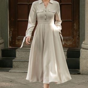 May include: A beige long-sleeved dress with a collared neckline, button closure, and pockets. The dress has a fitted bodice and a flowing skirt.
