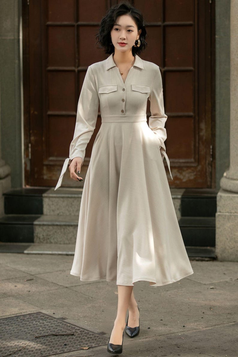 May include: A beige long-sleeved dress with a collared neckline, button closure, and pockets. The dress has a fitted bodice and a flowing skirt.
