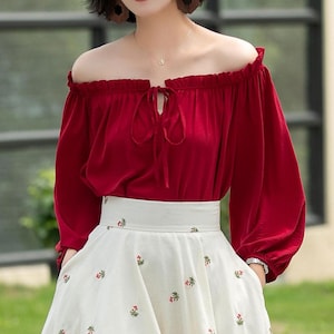 Könnte beinhalten: Eine rote, schulterfreie Bluse mit langen Ärmeln und einem Knoten am Ausschnitt. Die Bluse wird mit einem weißen Rock mit Blumenmuster kombiniert.