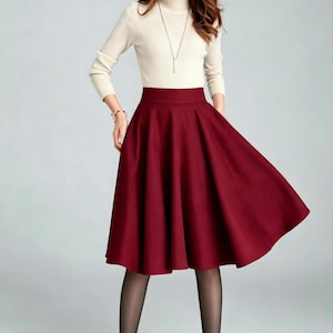 May include: A cream-colored long-sleeve mock neck top paired with a solid deep red midi-length skirt. The skirt features a high waistband and a flared, pleated silhouette that falls below the knees. The outfit is completed with sheer black tights.