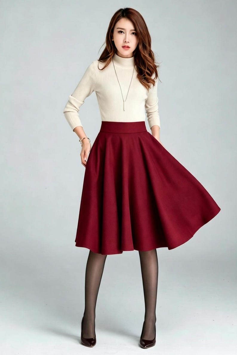 May include: A cream-colored long-sleeve mock neck top paired with a solid deep red midi-length skirt. The skirt features a high waistband and a flared, pleated silhouette that falls below the knees. The outfit is completed with sheer black tights.