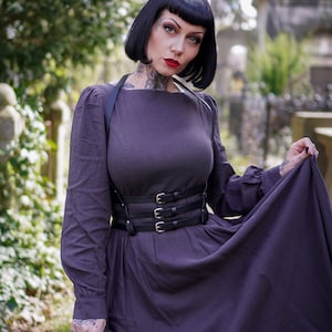 May include: A woman wearing a dark purple dress with a black leather harness and belt. The dress has a square neckline and long sleeves. She is standing in a cemetery setting.