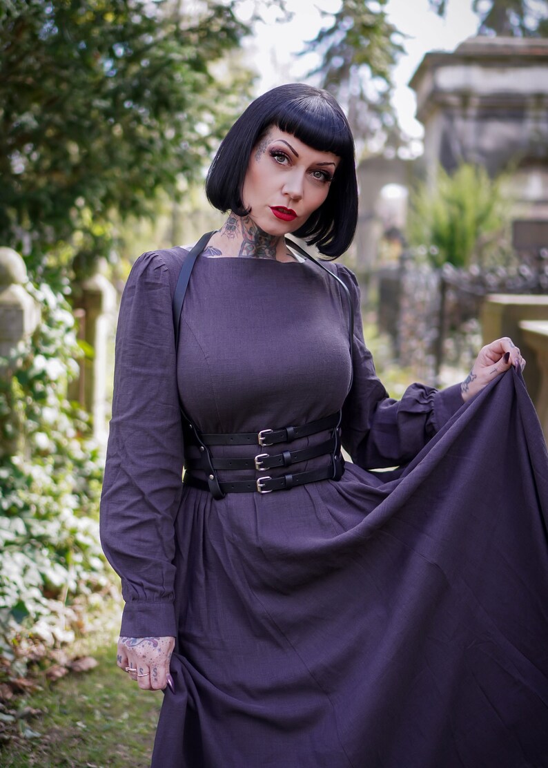 May include: A woman wearing a dark purple dress with a black leather harness and belt. The dress has a square neckline and long sleeves. She is standing in a cemetery setting.