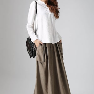 May include: A woman wearing a white long-sleeved shirt and a brown maxi skirt with pockets. She is carrying a black shoulder bag and wearing brown ankle boots.