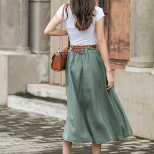 May include: A woman wearing a white t-shirt and a green linen skirt with a brown belt. She is carrying a brown leather handbag.