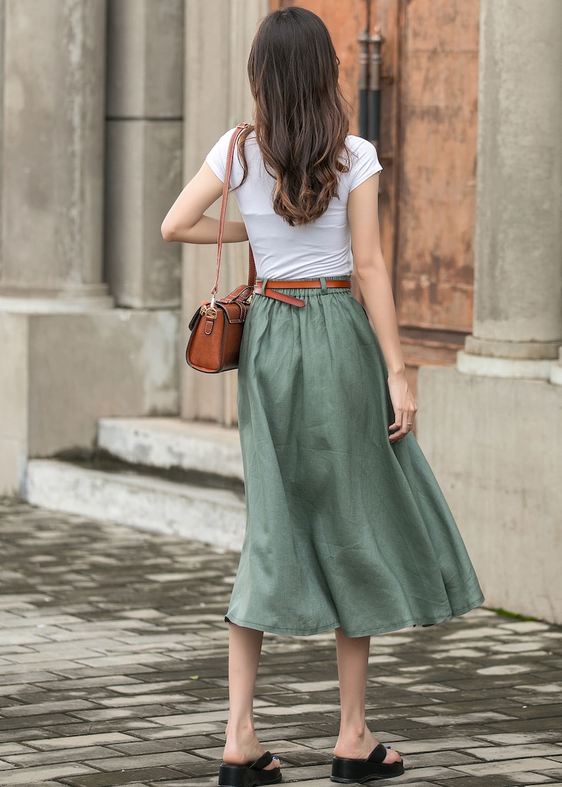 May include: A woman wearing a white t-shirt and a green linen skirt with a brown belt. She is carrying a brown leather handbag.