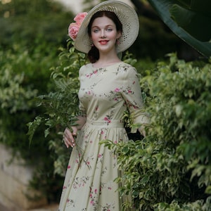 Peut inclure: Une femme porte une robe cr&egrave;me &agrave; imprim&eacute; floral et un chapeau de paille &agrave; larges bords orn&eacute; de roses roses. La robe a une taille ajust&eacute;e et des manches longues. Elle tient une branche de feuillage vert, devant une haie verte luxuriante.
