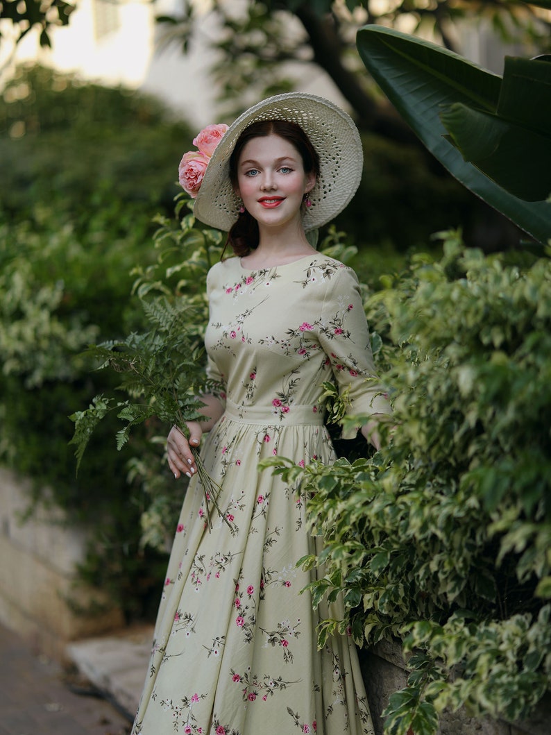 Peut inclure: Une femme porte une robe cr&egrave;me &agrave; imprim&eacute; floral et un chapeau de paille &agrave; larges bords orn&eacute; de roses roses. La robe a une taille ajust&eacute;e et des manches longues. Elle tient une branche de feuillage vert, devant une haie verte luxuriante.