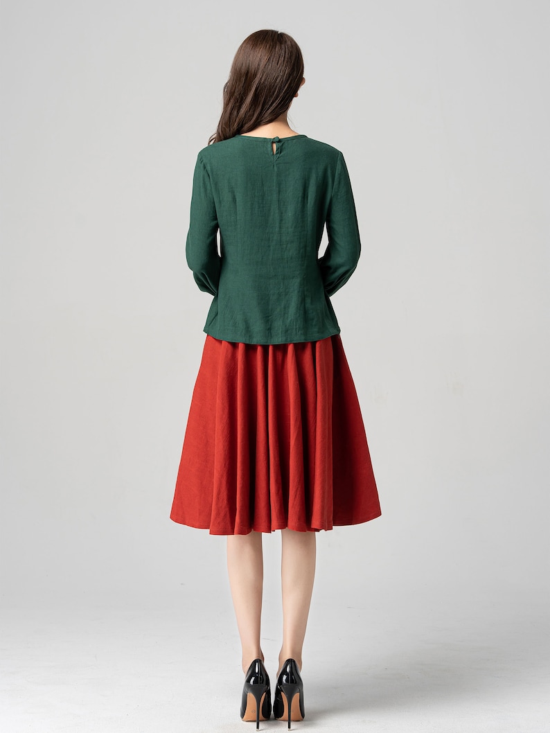 May include: A woman wearing a green long-sleeved blouse and a red midi skirt. She is wearing black high heels.