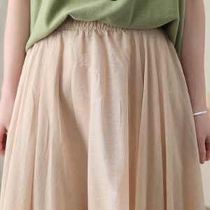 May include: A beige skirt with an elastic waistband and a flowing, pleated design. The skirt is paired with a green top. The fabric appears to be lightweight and suitable for warm weather.