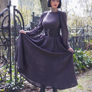 May include: A woman wearing a dark brown dress with a black leather corset belt and black boots. She is standing in front of a black metal fence.