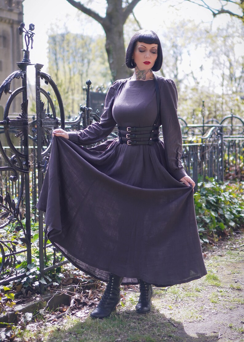 May include: A woman wearing a dark brown dress with a black leather corset belt and black boots. She is standing in front of a black metal fence.