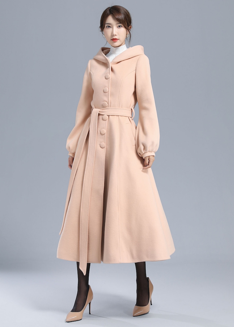 May include: A long, light pink coat with a hood, a belt, and button closures. The coat has a flared skirt and long sleeves.