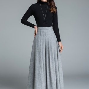 May include: A woman's outfit featuring a black long-sleeved turtleneck sweater and a gray pleated maxi skirt. The skirt falls to the ankles. The woman is wearing a silver necklace and black boots. The background is a solid gray.
