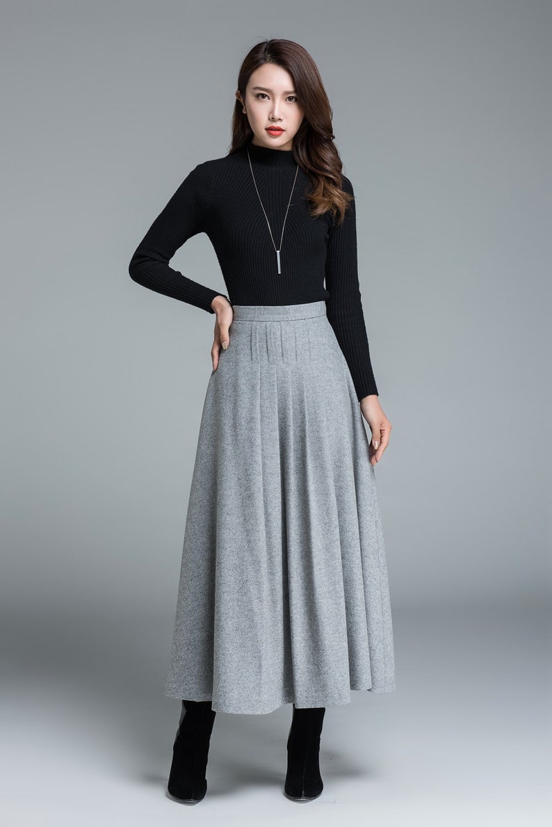 May include: A woman's outfit featuring a black long-sleeved turtleneck sweater and a gray pleated maxi skirt. The skirt falls to the ankles. The woman is wearing a silver necklace and black boots. The background is a solid gray.