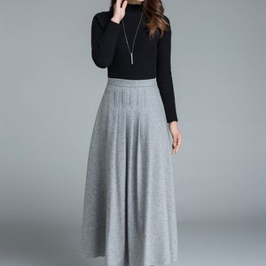 May include: A woman's outfit featuring a black turtleneck sweater and a long, gray pleated skirt. The skirt falls to the ankles, and the outfit is completed with black boots and a silver necklace. The background is a solid gray.