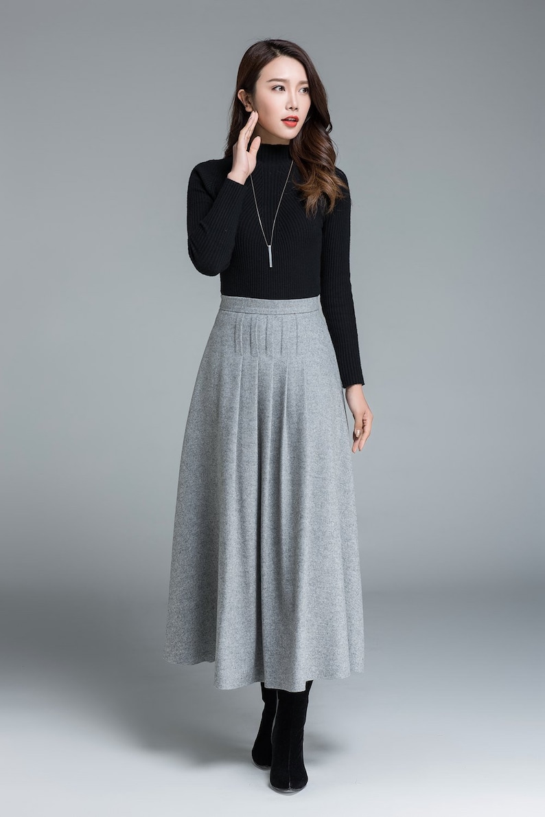 May include: A woman's outfit featuring a black turtleneck sweater and a long, gray pleated skirt. The skirt falls to the ankles, and the outfit is completed with black boots and a silver necklace. The background is a solid gray.