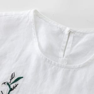 May include: Close-up of a white linen top with a round neckline and a small keyhole opening at the back. The top features an embroidered floral design in green and black on the front, suggesting a casual, everyday wear garment.