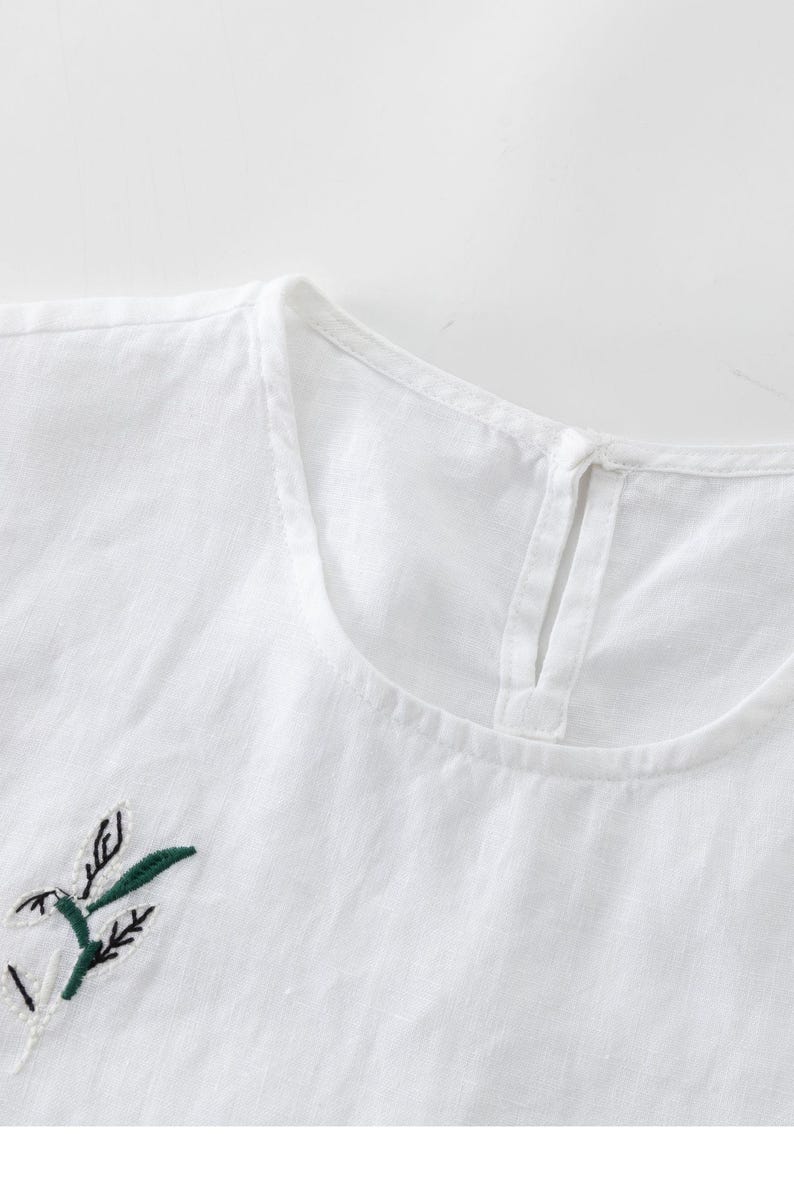 May include: Close-up of a white linen top with a round neckline and a small keyhole opening at the back. The top features an embroidered floral design in green and black on the front, suggesting a casual, everyday wear garment.