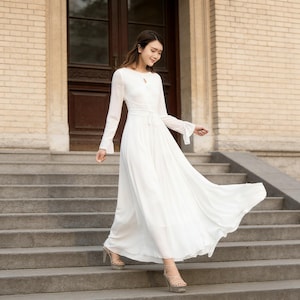 Puede incluir: Un vestido largo y blanco con mangas largas y escote de ojo de cerradura. El vestido presenta una cintura ce&ntilde;ida y una falda acampanada. La modelo lleva tacones de plataforma y baja una escalera de piedra.