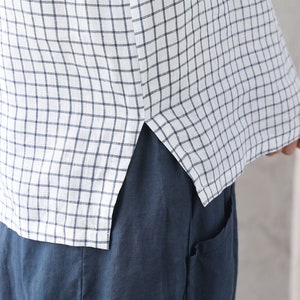 Op de afbeelding: Een wit linnen overhemd met een blauw-wit geruit patroon. Het overhemd heeft een zijsplit aan de onderkant en is gestopt in een blauwe linnen broek.