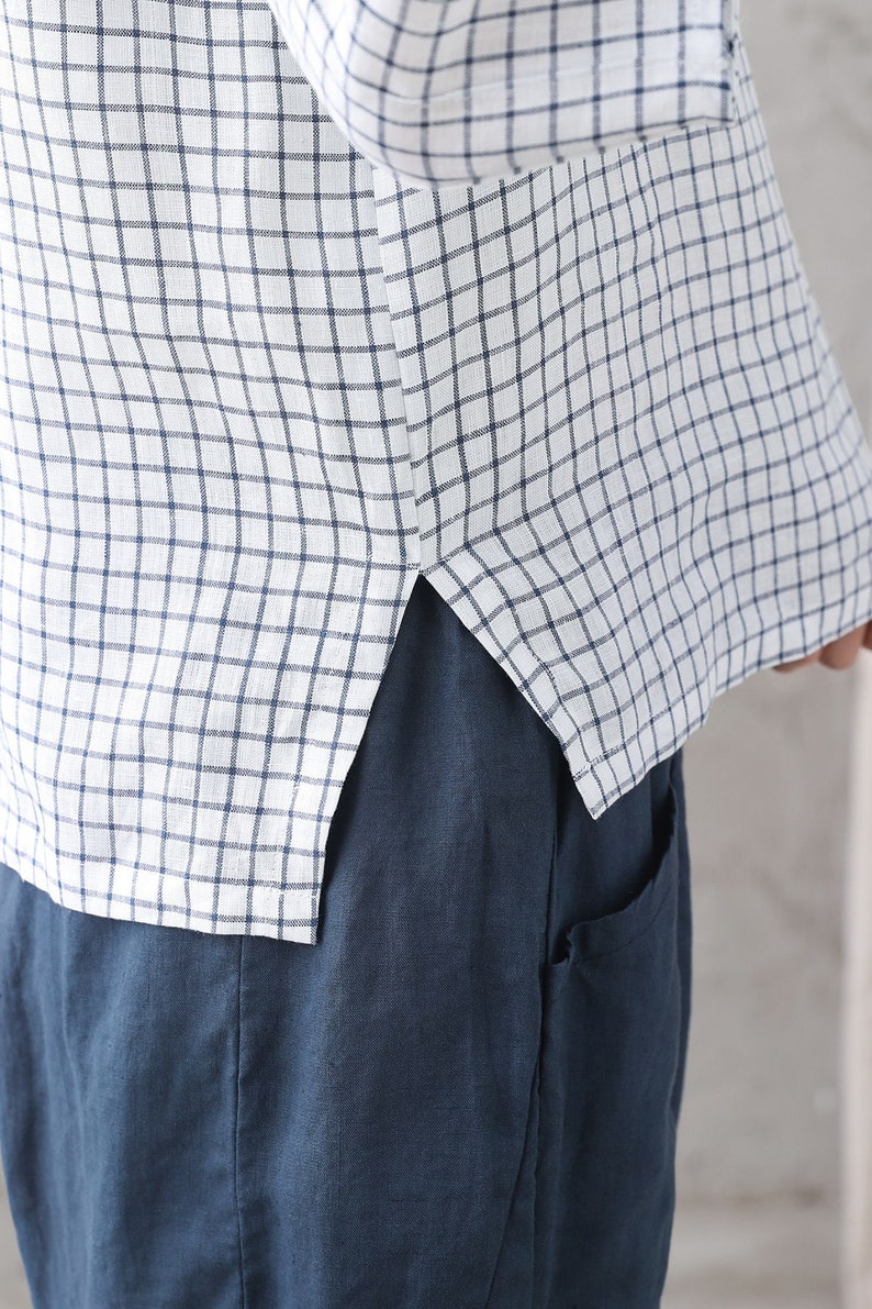 Op de afbeelding: Een wit linnen overhemd met een blauw-wit geruit patroon. Het overhemd heeft een zijsplit aan de onderkant en is gestopt in een blauwe linnen broek.