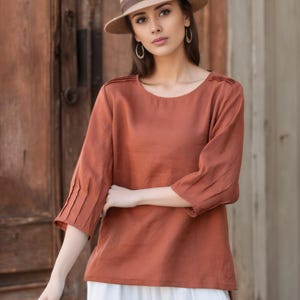 May include: A woman wearing a rust-colored linen top with pleated sleeves and a straw hat. She is standing in front of a wooden door.