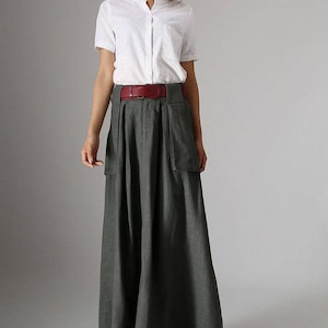 May include: A woman wearing a white collared shirt with a brown belt and a long, dark gray skirt with large pockets. She is wearing brown ankle boots.