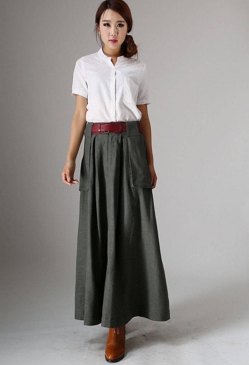 May include: A woman wearing a white collared shirt with a brown belt and a long, dark gray skirt with large pockets. She is wearing brown ankle boots.
