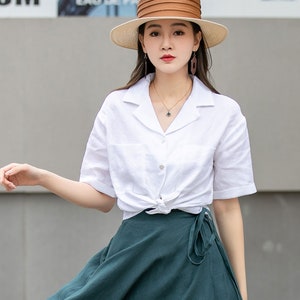 May include: A woman wearing a white linen shirt with a knotted front, a dark green wrap skirt, and a wide-brimmed straw hat with a brown leather band.