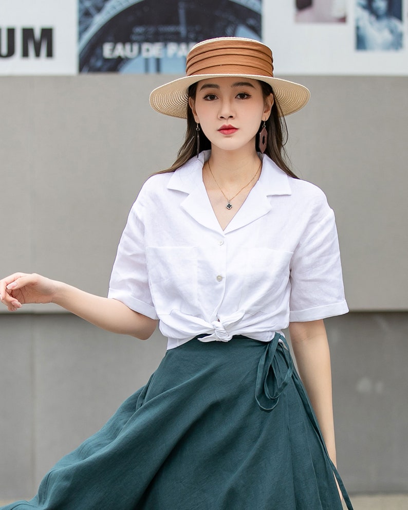 May include: A woman wearing a white linen shirt with a knotted front, a dark green wrap skirt, and a wide-brimmed straw hat with a brown leather band.