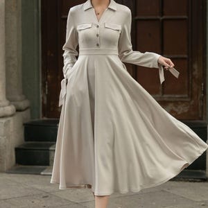 May include: A beige long-sleeved dress with a collared neckline, button closure, and a flared skirt. The dress has pockets on the sides and ties at the cuffs.