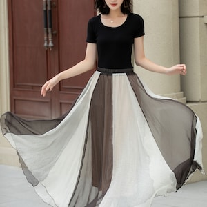 May include: A woman wearing a black top and a white hat is standing in front of a wooden door. She is wearing a long, flowing skirt with black, white and brown panels. The skirt is made of a sheer fabric and has a ruffled hem.
