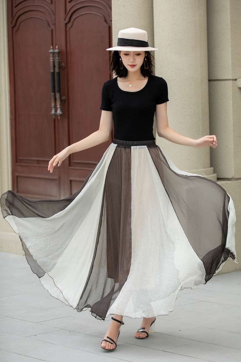 May include: A woman wearing a black top and a white hat is standing in front of a wooden door. She is wearing a long, flowing skirt with black, white and brown panels. The skirt is made of a sheer fabric and has a ruffled hem.