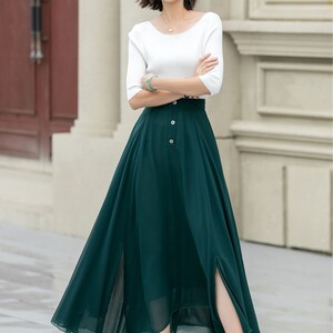 May include: A woman wearing a white top and a long, dark green skirt with a button closure and a high slit. The skirt is made of a flowy fabric and has a slightly asymmetrical hemline.