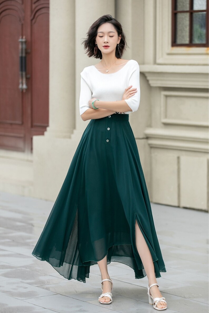 May include: A woman wearing a white top and a long, dark green skirt with a button closure and a high slit. The skirt is made of a flowy fabric and has a slightly asymmetrical hemline.