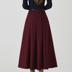 May include: A woman wearing a black long-sleeved top and a burgundy midi skirt. The skirt has a high waist and pleats, and is paired with a black belt. The woman is also wearing black heels.