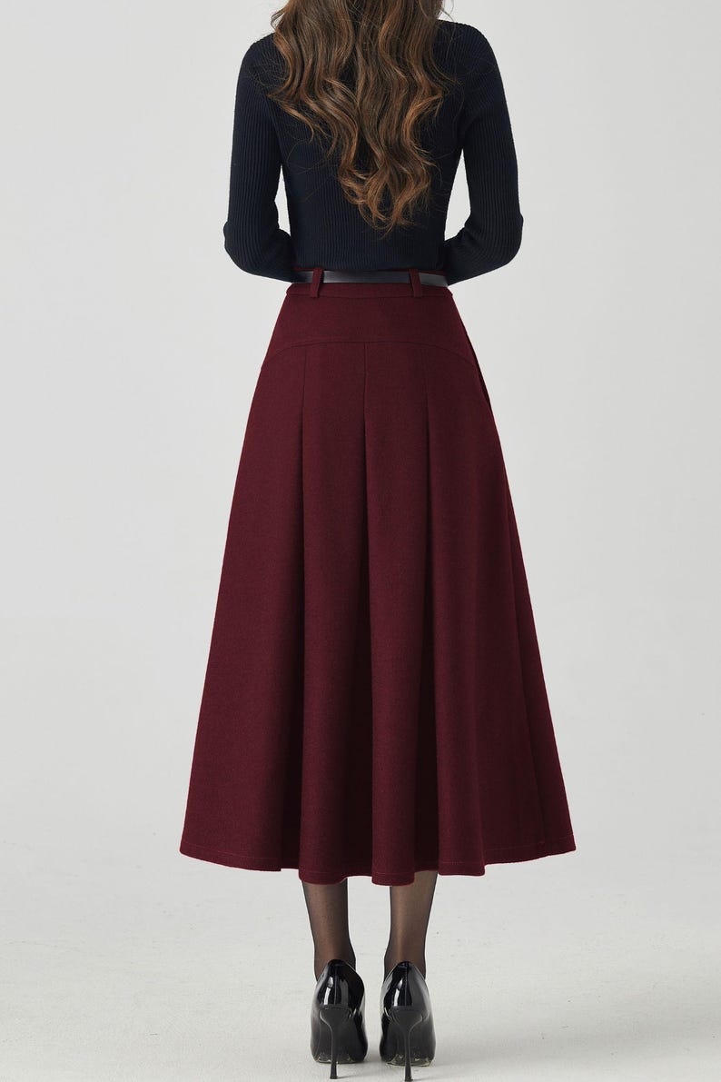 May include: A woman wearing a black long-sleeved top and a burgundy midi skirt. The skirt has a high waist and pleats, and is paired with a black belt. The woman is also wearing black heels.