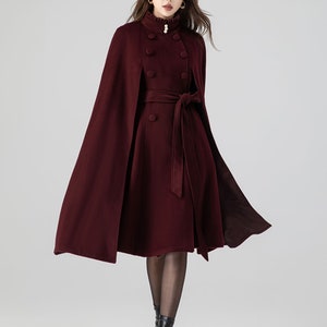 Pu&ograve; includere: Un cappotto a mantella color bordeaux intenso con collo alto a volant e un bottone perlato, bottoni rivestiti in tessuto a doppio petto e una cintura in vita abbinata. Il capo presenta una silhouette svasata che arriva al ginocchio.