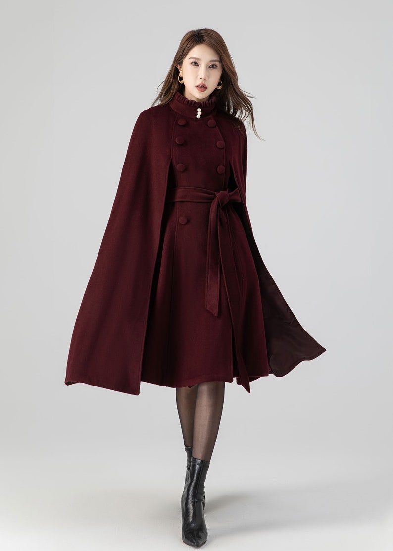 Pu&ograve; includere: Un cappotto a mantella color bordeaux intenso con collo alto a volant e un bottone perlato, bottoni rivestiti in tessuto a doppio petto e una cintura in vita abbinata. Il capo presenta una silhouette svasata che arriva al ginocchio.