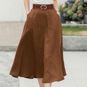 May include: A brown linen midi skirt with a brown leather belt and a gold buckle. The skirt has a slight A-line shape and falls to just below the knee.