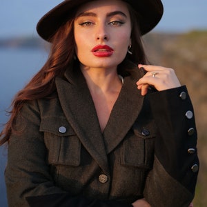 Puede incluir: Una mujer con una chaqueta verde oscuro con ribetes y botones negros. Lleva un sombrero fedora marr&oacute;n y tiene los labios pintados de rojo.