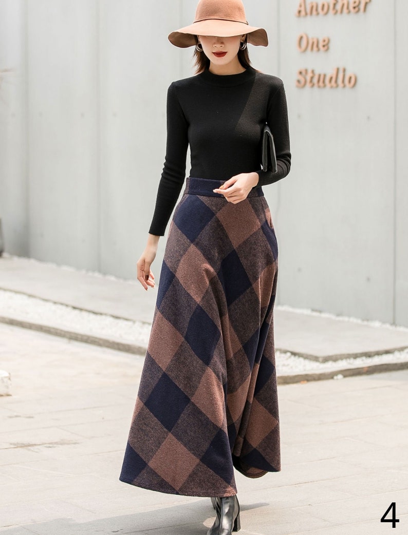 May include: A woman wearing a black long-sleeved shirt and a brown and blue plaid skirt. She is wearing a brown hat and black boots. The skirt is a maxi skirt and falls to her ankles.