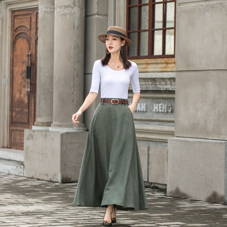 May include: A woman wearing a white, scoop-neck, three-quarter sleeve top, a long olive-green skirt, and a brown belt. She is also wearing a straw hat and heels. The outfit is suitable for a casual or semi-formal occasion.