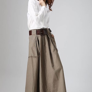 May include: A brown linen skirt with a wide brown leather belt and a large buckle. The skirt has a pleated front and two large pockets on the sides. The model is wearing a white long-sleeved shirt.