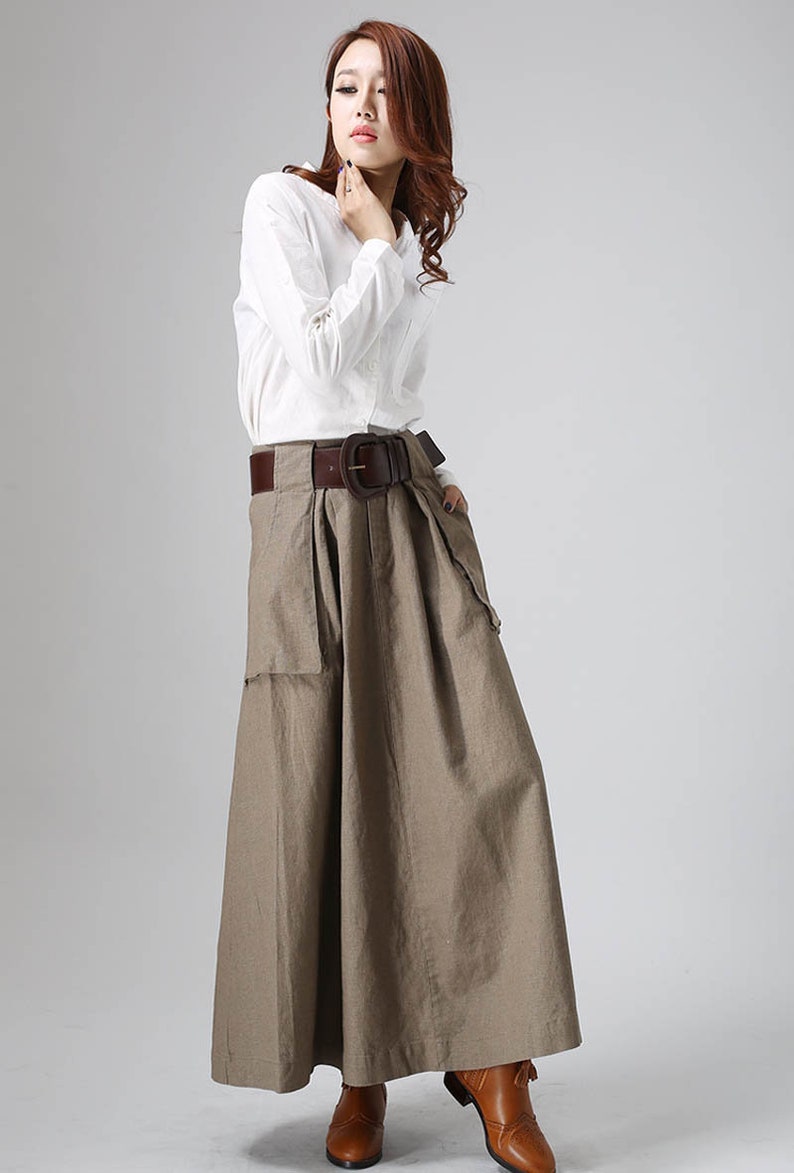 May include: A brown linen skirt with a wide brown leather belt and a large buckle. The skirt has a pleated front and two large pockets on the sides. The model is wearing a white long-sleeved shirt.
