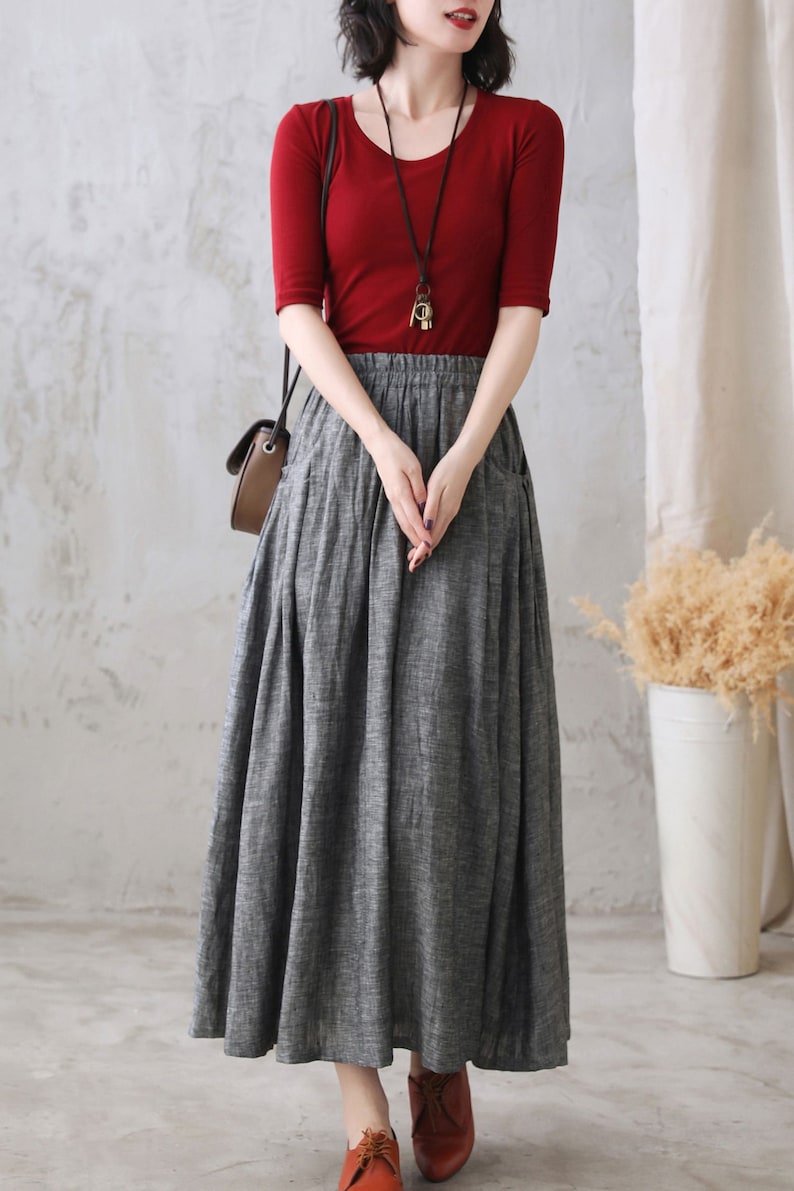 May include: A gray linen skirt with a red top and a brown leather bag. The skirt has a high waist and a flowy silhouette. The top is a simple, fitted style with short sleeves. The bag is a small, crossbody style.